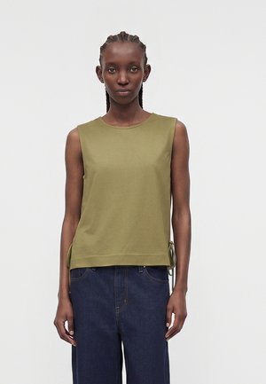Femme aux cheveux tressés portant un haut sans manches vert olive et un jean taille haute bleu foncé, debout devant un fond clair uni.