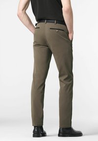 Man in olijfgroene slim-fit broek, zwarte gevlochten riem, zwart shirt en zwarte leren Chelsea-boots, staande met handen in de zakken.