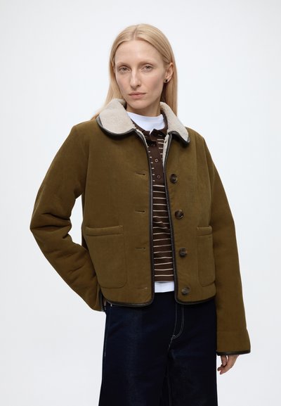 Femme aux longs cheveux blonds portant une veste marron boutonnée avec un col en peau lainée claire par-dessus une chemise rayée et un pantalon foncé.