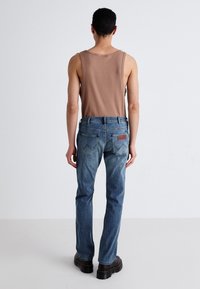 Jeans de mezclilla azul para hombre con un corte recto, que cuentan con bolsillos traseros, un parche de cuero marrón y un ligero desvanecimiento. Combinados con una camiseta sin mangas beige.
