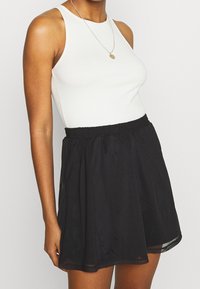 Mujer vistiendo una blusa sin mangas blanca y una falda negra de cintura alta con banda elástica, accessorada con un collar de colgante redondo.