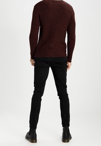 Man wearing a brown textured long-sleeve sweater, black slim pants, and black ankle boots standing with back facing the viewer.