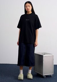 Camiseta negra de gran tamaño con texto bordado, combinada con pantalones de denims oscuros de pierna ancha y zuecos verde claro, al lado de un estuche metálico plateado.