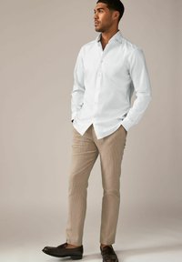 Hvid skjorte med knapper, beige teksturerede bukser og mørkebrune sko båret af en mandlig figur. Stående positur med hænderne i lommerne.