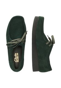 Vihreät mokkasiinimalliset kengät, joissa on pyöreä kärki, nauhakiinnitys ja musta teksturoitu kumipohja.