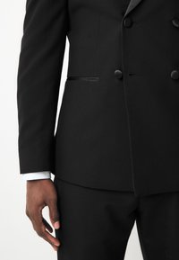 Black tuxedo with textured fabric, double-breasted design, rounded black buttons, and a slim fit. White shirt cuffs visible at the wrist.