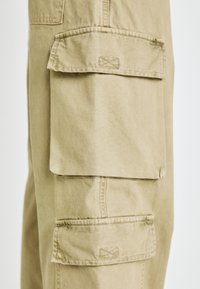 Beige Cargo-Hosen mit mehreren großen Taschen mit Klappen, genähten Details und einer glatten Baumwolltextur.