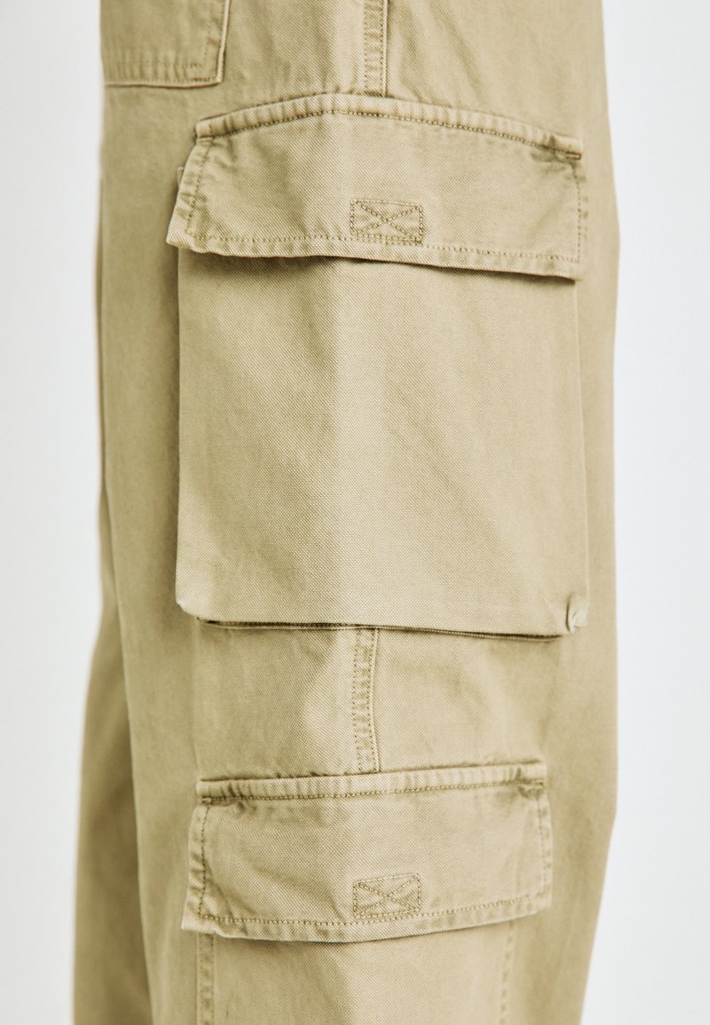 Pantalones cargo beige con múltiples bolsillos grandes con solapas, detalles de costura y una textura de algodón suave.