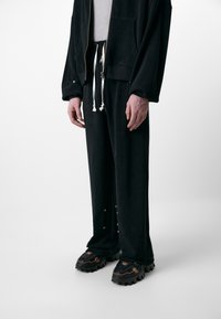 Black fleece tracksuit with a zip-up jacket, elastic waistband, and drawstrings. Includes large pockets and snap details on pants. Chunky black sneakers.