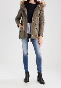 Olivgrüner Parka mit gefütterter Kapuze, zwei vorderen Taschen und Zugband in der Taille. Getragen über einem dunklen Oberteil, kombiniert mit gradienten Jeans und schwarzen Stiefeln.
