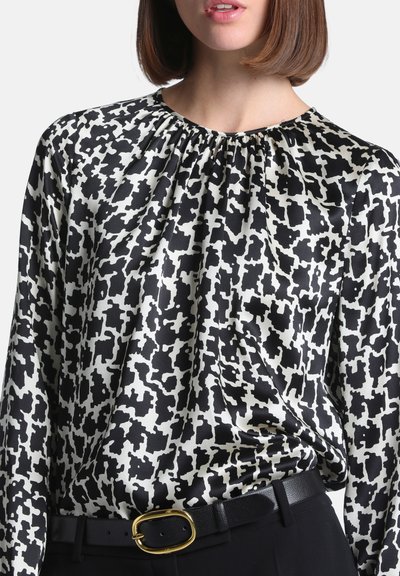 Femme portant un chemisier à motif abstrait noir et blanc rentré dans un pantalon noir avec une ceinture noire ornée d'une boucle ovale dorée.