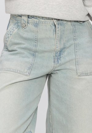 Jean en denim bleu clair taille haute avec grandes poches avant patch et détails de coutures visibles, porté avec un haut gris clair.