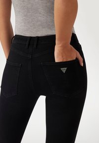 Los jeans de mezclilla negra tienen un diseño ajustado, dos bolsillos traseros y un pequeño logo de la marca en forma de triángulo en uno de los bolsillos. La cintura es elástica.