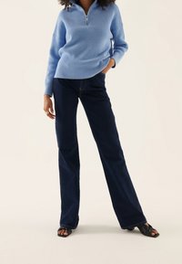 Model trägt einen hellblauen, gerippten Pullover mit Reißverschluss und dunkle Jeans, steht vor einem neutralen Hintergrund. Stylische Schuhe vervollständigen den Look.