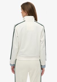 Witte jas met ritssluiting, donkere groene zijstrepen, hoge kraag, geribbelde boorden en zoom. Gladde textuur, ontworpen voor casual gebruik.