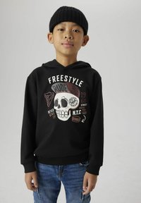 Hoodie noir avec un design graphique représentant un crâne et divers éléments textuels. Porté avec un jean bleu. Poignets et ourlet côtelés. Matière en coton.