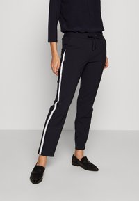 Marineblauwe broek met elastische taille, witte zijkanten, rechte pasvorm en trekkoord. Gecombineerd met zwarte loafers. Soepele textuur, casual ontwerp.