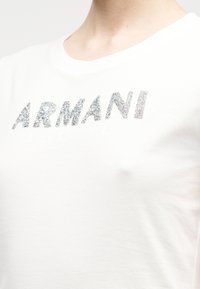 T-shirt branca de mangas curtas com logotipo "ARMANI" prateado brilhante e texto mate "EXCHANGE" abaixo. Tecido macio de algodão com ligeira textura.