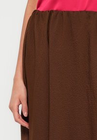 Brązowa spódnica o fakturze z marszczonym paskiem w talii, noszona z różowym topem. Tkanina ma subtelny prążkowany wzór.