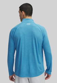 Lichtblauw sportshirt met lange mouwen, een gestructureerd patroon, kraag en logo op de achterkant. Heeft raglanmouwen voor verbeterde mobiliteit.