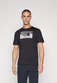 Mees, kes kannab musta The North Face t-särki, millel on topograafia kujundus ja loosung "Ära peatu uurimisel", seisab ühtlasel taustal.