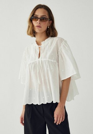 Kobieta z krótkimi brązowymi włosami, brązowymi owalnymi okularami przeciwsłonecznymi, luźną białą bluzką z plisami i falowanym dołem oraz czarnymi spodniami na jednolitym tle.