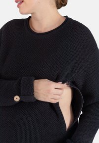 Pull noir en texture avec un col rond et des manches longues. Dispose d'une fermeture éclair sur le côté, permettant d'accéder à la couche inférieure. Détail de bouton en bois.
