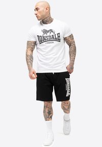 Fehér póló fekete "Lonsdale London" logóval, fekete sportnadrág fehér logóval az oldalán, fehér sportcipővel és fehér zoknival viselve.