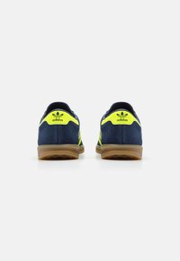 Tmavě modré sportovní semišové boty se žlutými akcenty, s gumovou podrážkou a klasickým třímásým vzorem. Pohled zezadu ukazuje značku.