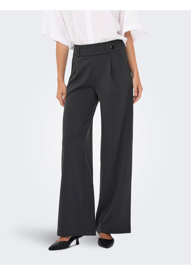 Femme portant un pantalon noir taille haute à jambes larges avec des détails de boutons, associé à une chemise blanche et des chaussures noires à talons.