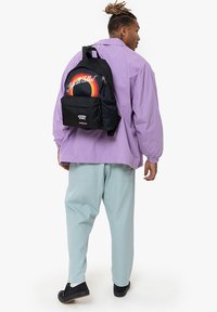 Sac à dos noir avec un design coloré incurvé et le texte "C'est tout, mes amis !" ainsi qu'une poche avant. Porté par-dessus une veste violette.