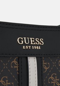 Black leather wallet with gold "GUESS EST 1981" logo, featuring a textured pattern in tan and white accents. Smooth finish with stitching details.