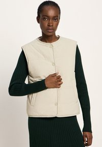 Hellbeige gesteppte Weste mit Knopfverschluss, mit hohem Kragen und ärmellosem Design, über einem dunkelgrünen gerippten Pulloverdress getragen.