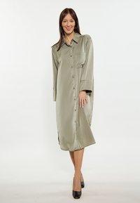Olivegrünes Satin Hemdkleid, lange Ärmel, Knopfverschluss vorne, seitliche Schlitze am Saum, eine Brusttasche, kombiniert mit schwarzen Schuhen.