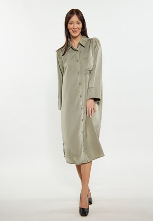 Olivegrünes Satin Hemdkleid, lange Ärmel, Knopfverschluss vorne, seitliche Schlitze am Saum, eine Brusttasche, kombiniert mit schwarzen Schuhen.