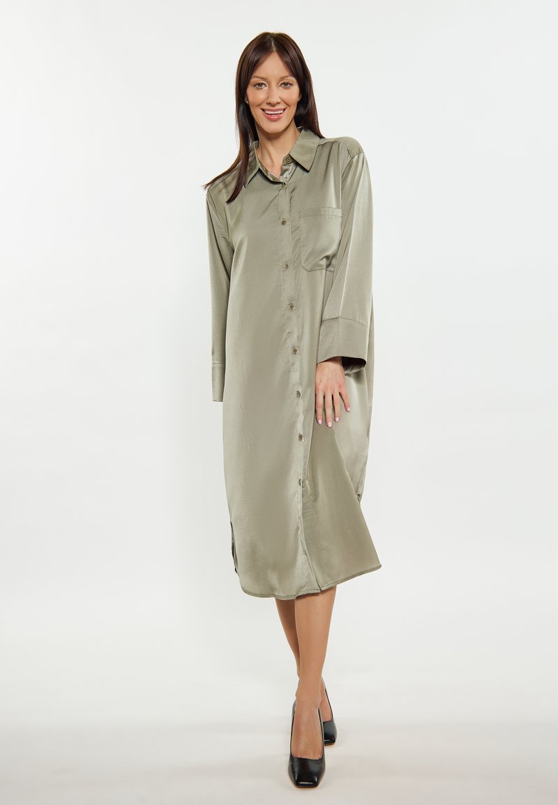 Olivegrünes Satin Hemdkleid, lange Ärmel, Knopfverschluss vorne, seitliche Schlitze am Saum, eine Brusttasche, kombiniert mit schwarzen Schuhen.
