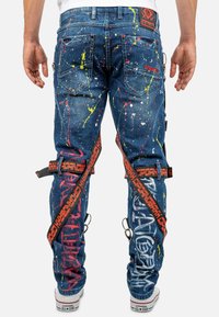 Jean en denim bleu avec un design de tache de peinture jaune, rose et blanc. Caractéristiques des bretelles rouges avec logo et accents en métal.