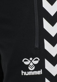 Svart tyg med en vertikal svart dragkedja, vita chevronränder och en vit stiliserad bi-logotyp ovanför ordet "hummel" i små bokstäver.