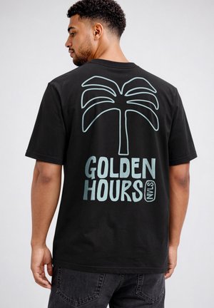 Man with short curly hair wearing a black t-shirt featuring a large light blue palm tree and "GOLDEN HOURS NVLS" text on the back.