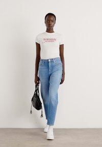 Bela majica s rdečim logotipom "Tommy Jeans", svetlo modri naravnost krojeni kavbojki, črna torba in bele superge. Model stoji proti svetli podlagi.