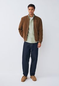 Giacca bomber in suede marrone, camicia verde chiaro con bottoni, jeans a gamba larga blu scuro e scarpe in suede color cuoio. Descrizione di un outfit casual a strati.