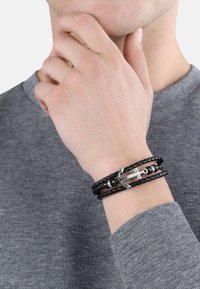 Bracciale intrecciato in pelle nera con chiusura ad ancora in argento e dettagli cilindrici, indossato su un polso sopra un maglione grigio texturizzato.