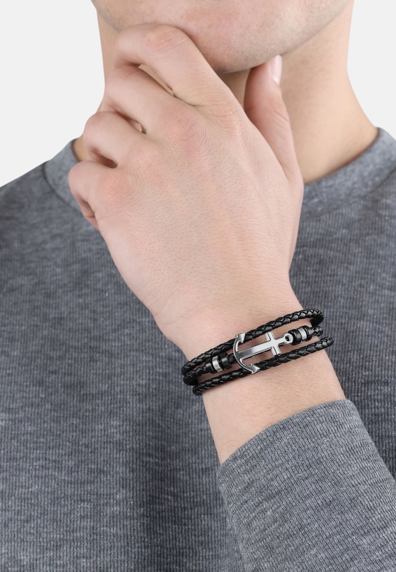 Bracciale intrecciato in pelle nera con chiusura ad ancora in argento e dettagli cilindrici, indossato su un polso sopra un maglione grigio texturizzato.
