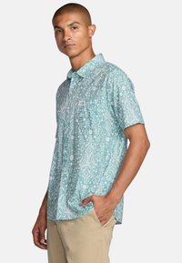 Short-sleeve button-up shirt in light teal with a white abstract pattern; features a chest pocket and a brand label on the front.