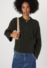 Dark green knitted jacket with a high collar, front zipper, and two front pockets. Material is textured and slightly fuzzy.