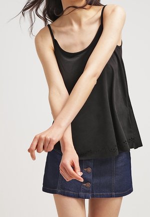 Kvinde iført en sort ærmeløs top med tynde stropper og en blå denim mini-nederdel med knapper foran, stående med armene over kors.