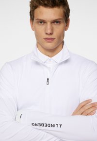 Valkoinen pitkähihaisella pulloverilla, jossa on vetoketjukaulus ja logotiedot hihassa. Tasainen pinta ja istuva muotoilu.