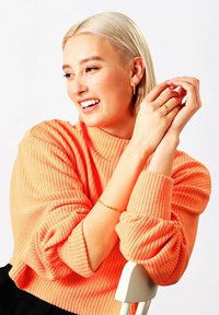 Maglione arancione a coste con collo alto, abbinato a orecchini a cerchio dorati, un sottile bracciale d'oro e un anello. Modella seduta su una sedia.