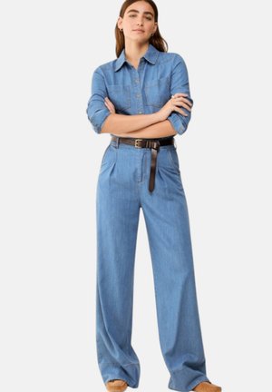 Donna con lunghi capelli castani, che indossa una camicia di jeans blu con bottoni infilata in pantaloni di jeans a vita alta e gamba larga con una cintura scura, in piedi con le braccia incrociate.