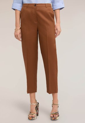 Vrouw draagt cropped bruine broek, blauwe opgerolde mouwen van een overhemd en patroonhooggehakte sandalen, staat met ontspannen handen langs het lichaam.